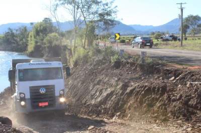 Caminhões estão colocando pedras detonadas para conter erosão da margem do rio sobre a rodovia